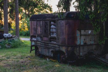 Meadow 'da büyük bir ladin altında park edilmiş eski model bir karavan. Arka planda eski çatı, yeşillik ve gökyüzü.