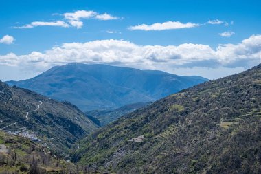 Kayalık dağların dibi kısmen karla kaplı arka planda açık mavi gökyüzü ve ön planda kısmen büyümüş kil uçurum boyunca boş asfalt yol..