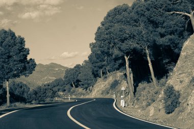 Resimli kırmızı kil kayalıklar boyunca boş dolambaçlı yol kısmen ağaçlarla ve mavi gökyüzü ve arka planda beyaz bulutlarla kaplı çalılarla kaplı. Güneşli bir yaz günü.