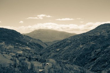 Kayalık dağlara bak. Kısmen karla kaplı. Arka planda bulutlar ve ön planda yemyeşil ağaçlar.