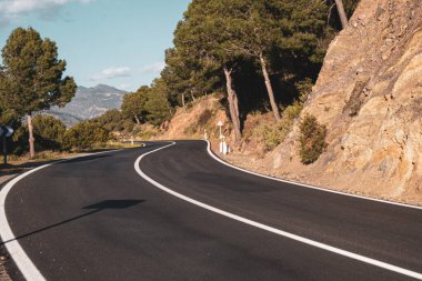 Resimli kırmızı kil kayalıklar boyunca boş dolambaçlı yol kısmen ağaçlarla ve mavi gökyüzü ve arka planda beyaz bulutlarla kaplı çalılarla kaplı. Güneşli bir yaz günü.