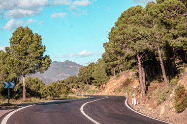 Resimli kırmızı kil kayalıklar boyunca boş dolambaçlı yol kısmen ağaçlarla ve mavi gökyüzü ve arka planda beyaz bulutlarla kaplı çalılarla kaplı. Güneşli bir yaz günü.