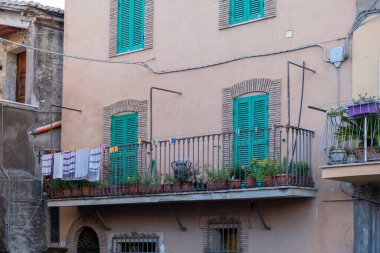 Etkileyici turkuaz boyalı pencere panjurlarıyla binaya bakın. Metal dizayn edilmiş balkon. Kuruması için giysi asılmış. Çiçek saksısı. Binanın duvarında elektrik kabloları var..