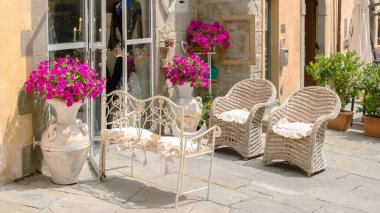 Açık camlı ön kapıya yerleştirilmiş klasik beyaz metal bank. Hasarlı alçılı sarımsı bina duvarı ve fuşya çiçekleri arka planda