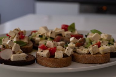 İtalyan ekmeği, Yunan peyniri, domates, salatalık, turp ve fesleğen ve çeşitli baharatlarla beyaz bir tabak ve beyaz bir mutfak masasında..
