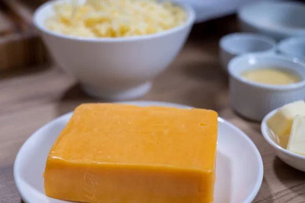 A piece of cheddar cheese on a white plate in the kitchen on a wooden table and white bowls with food ingredients on a blurred background. food background. Healthy food advertisement, poster for the kitchen. Space for your text