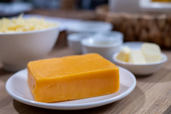 A piece of cheddar cheese on a white plate in the kitchen on a wooden table and white bowls with food ingredients on a blurred background. food background. Healthy food advertisement, poster for the kitchen. Space for your text