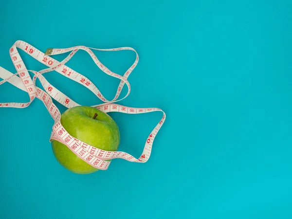 Green apple with measuring tape on blue background. The concept of weight loss, diet and nutrition intake tracking. Copy space.
