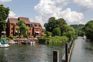 Marlow, Buckinghamshire 'daki Thames nehrine bakan Riverside apartmanları güneşli bir yaz öğleden sonrasında. Zevkli tekneler kıyıya demirledi ve diğerleri Maidenhead 'e doğru nehrin aşağısına doğru devam ediyor..