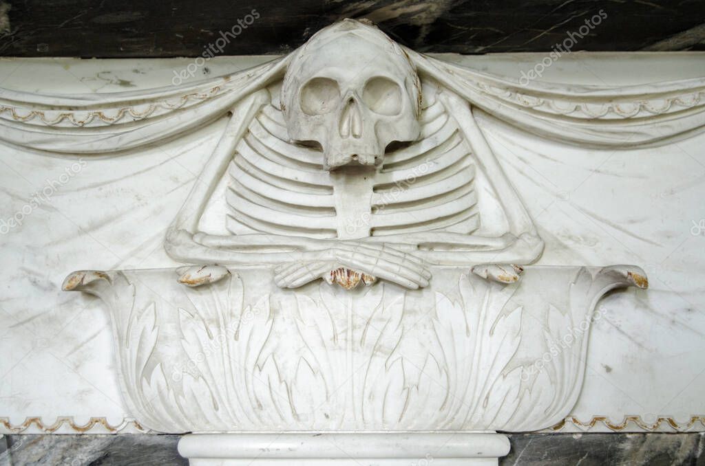 A marble carving from the 18th century showing a skeleton resting his folded arms on a plinth decorated with acanthus leaves.  On a memorial plaque which has been on display for over 150 years.