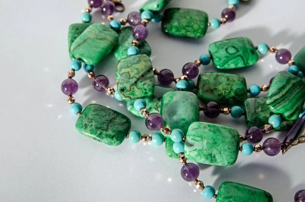 Coils of a handcrafted necklace made with beads of purple amethyst, blue turquoise and green crazy agate beads. Viewed on a white background with drop shadow.