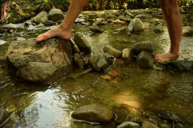Çıplak ayaklı bir kadının ormanda doğal bir derede yürürken bir kayaya doğru bir adım atması. Ayağında taşlar ve kayalarla.