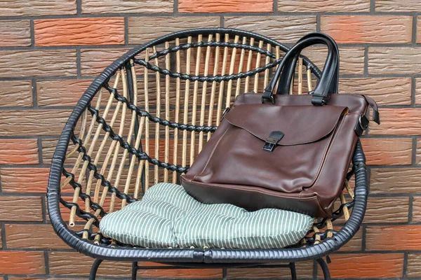 Classic brown leather bag left on wicker chair outdoors, background of brick wall.