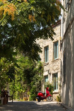 Meşhur, turistik Ege kasabası Alacati 'deki kırmızı bir motosiklet, ağaçlar ve eski, tarihi, geleneksel taş evler. Bu, Cesme ilinin bir köyüdür. Güneşli bir yaz günü.