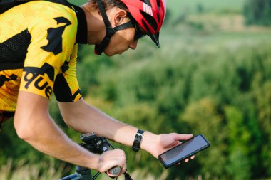 Happy male cyclist sending a text message on his mobile phone. Image of a cyclist on a mountain bike using a smartphone navigator exploring a map and searching for GPS coordinates while cycling.
