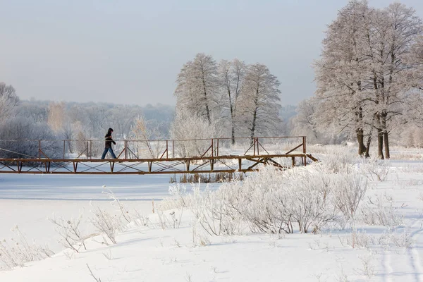 A man walks on a bridge over a river in winter. The frozen river is covered with ice and snow. Snow-covered footbridge across the river, winter landscape with trees and snow, frost on the trees.