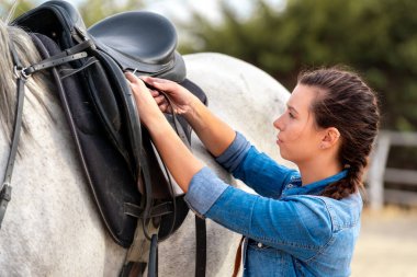 Side view of a young girl putting a saddle on a white horse in a stable. High quality photo
