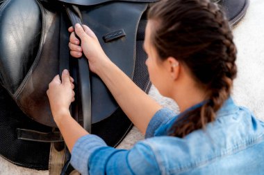 Side view of an unrecognizable young girl putting a saddle on a white horse in a stable. High quality photo
