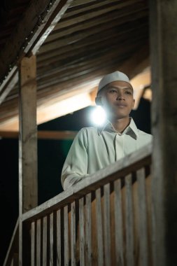 Karanlık bir gecede köyde beyaz bir Müslüman elbisesi giyerken balkonda dua eden İslami bir adam.