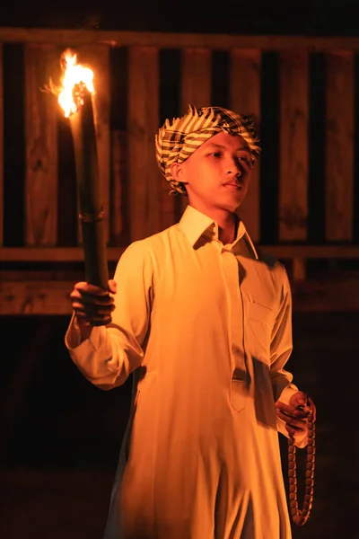 An Arabian man holding a fire torch in the front of a wooden house in the dark night