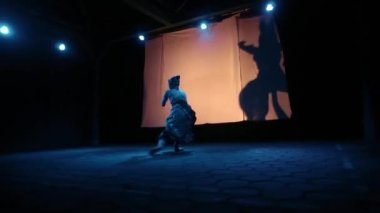 Mavi ışığın altında eski bir kostüm giyerken arka planda siluetle dans eden Asyalı kadın.