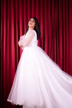 Güzel Asyalı kadın beyaz gelinlik içinde mutlu bir yüzle stüdyonun içinde kırmızı perdenin önünde duruyor.