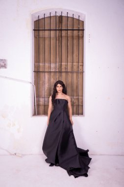 Beyaz bir stüdyoda, kırsal ahşap pencerenin önünde duran siyah elbiseli zarif kadın.