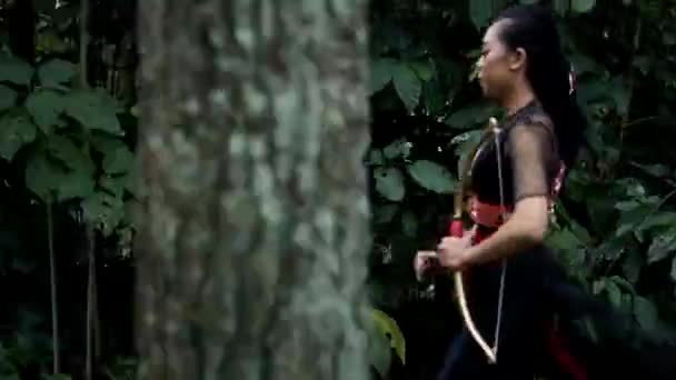 Ázsiai nők futnak gyorsan a dzsungelben, miközben egy nyilat és egy íjat tartanak a kezükben, és fekete jelmezt viselnek a testükön.