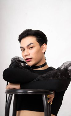 Siyah kostüm ve makyajla beyaz stüdyoda sandalyeyle poz veren yakışıklı Asyalı adam.