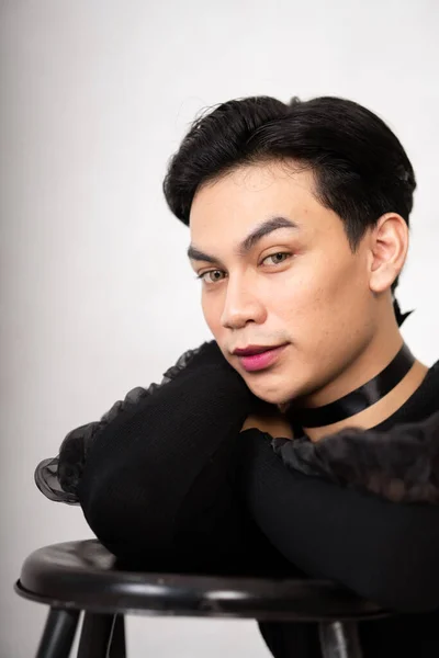 beautiful Asian man posing with the chair while wearing a black costume and makeup inside the white studio