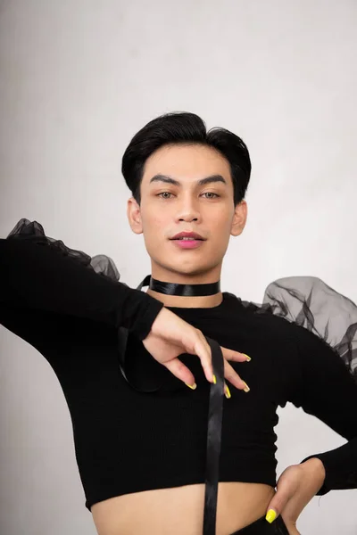 A transgender man poses in a black dress with an elegance face and black hair inside the white studio