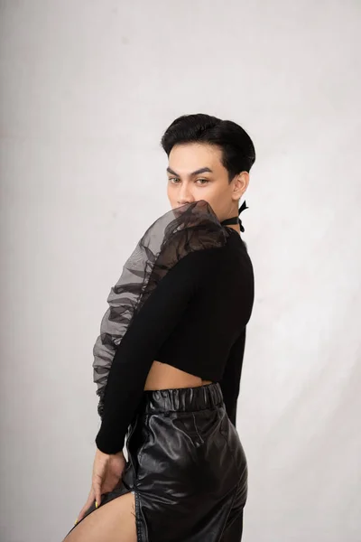 A transgender man poses in a black dress with an elegance face and black hair inside the white studio