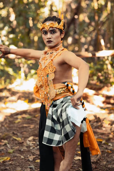 An angry Asian man poses shirtless while wearing a gold crown with stripped pants inside the forest