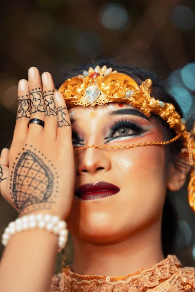 Asian woman with makeup covering his face with her hand while in makeup and wearing a gold crown and gold tiara during the daylight