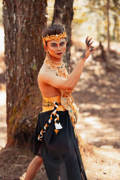 Manly Asian man posing in a golden crown and golden clothes while wearing makeup inside the forest