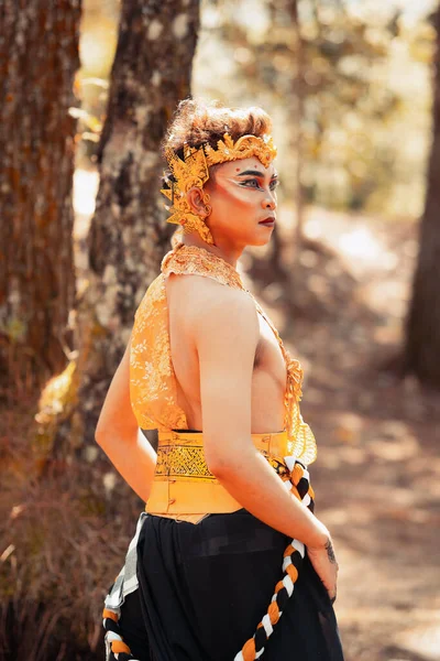 Manly Asian man posing in a golden crown and golden clothes while wearing makeup inside the forest