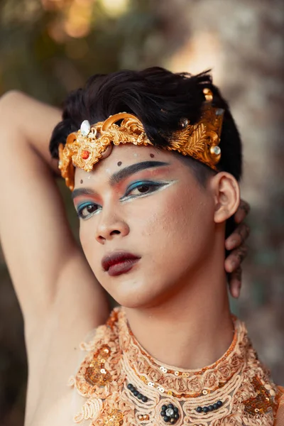 Sexy Asian man with a golden headpiece and golden necklace while in makeup near the beach in Bali