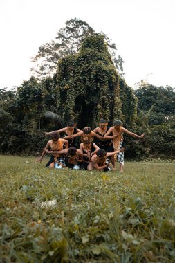 Bir grup Endonezyalı, tropikal ormandaki yeşil çimlerin üzerinde altın bir takım elbiseyle dans ederken poz veriyor.