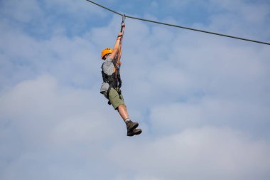 İp inerken ipe binen çocuk mavi bulutlu arka planda turuncu miğfer takıyor.