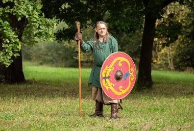 Yeşil giyinmiş Viking adam uzun baltayla ayakta duruyor ve elinde kırmızı tahta Viking kalkanı var.