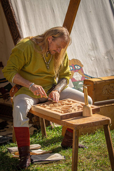 Viking carpenter with long hair doing woodcarving in front of tent during historic reenactment