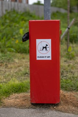 Köpek kakası torbaları için kırmızı çöp kutusu ve üzerinde köpek resmi olan beyaz çıkartma ve halka açık bir parkta temizleyin yazan kelimeler.