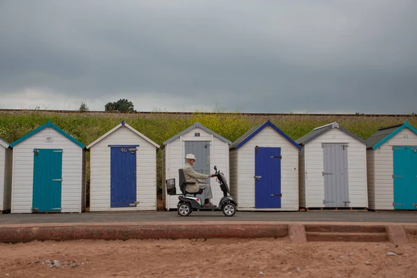 Old man on mobility scooter in front of beach huts during a cloudy summer afternoon
