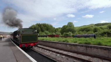 Steam train slowly driving along platform with smoke coming out of chimney at Bolton Abbey train station. Yorkshire Dales National Park