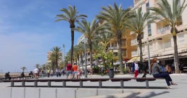 People walking and sitting on a bench at a popular tourist promenade in Torrevieja at Costa Blanca in Spain with palm trees waving in the wind