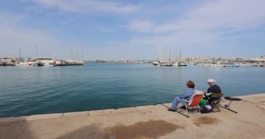 Two seniors sitting on a chair and fishing in the marina by the water with boats in the background