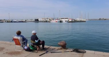 Two seniors enjoying time sitting on a chair and fishing by the water with boats in the background