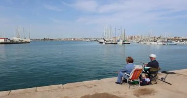 Two seniors sitting relaxing on a chair and fishing in the marina by the water with boats in the background
