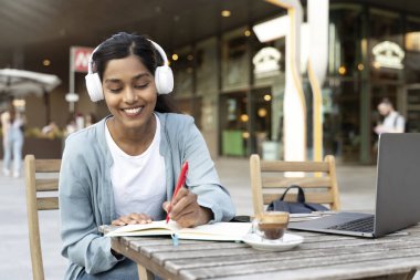 Authentic portrait of smiling Indian student wearing wireless headphones using laptop computer studying, taking notes, exam preparation, online education concept