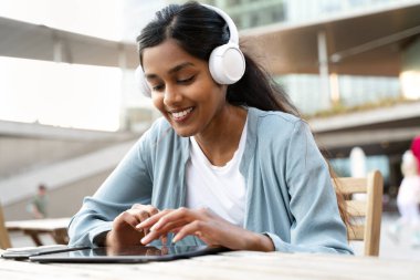 Dijital tablet kullanan, kablosuz kulaklık takan, video izleyen, modern kafede iletişim kuran gülümseyen Hintli kadın. Sosyal medya kavramı  